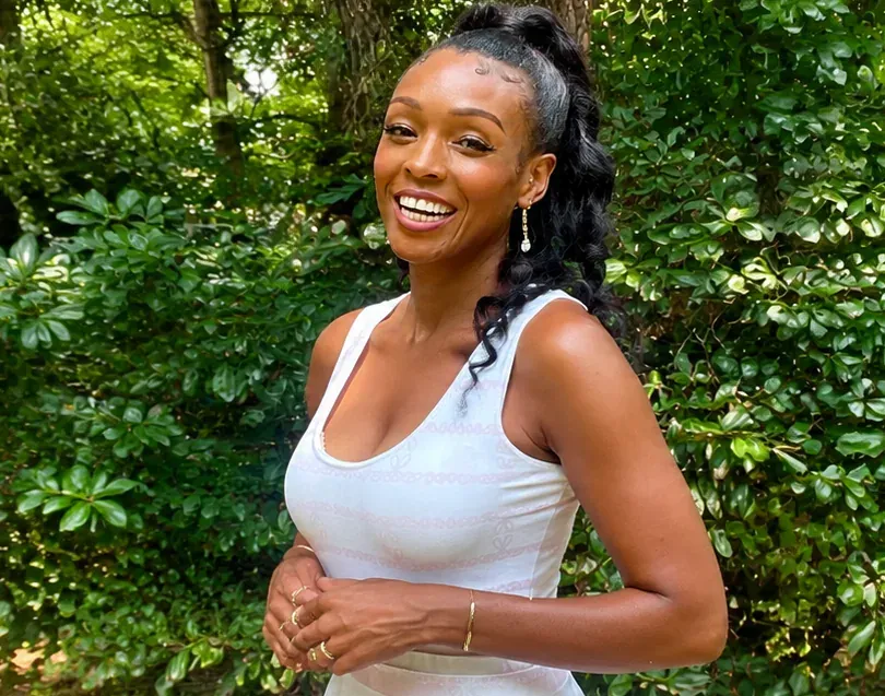 Ten Elyse smiling in a white dress in front of lush green garden foliage